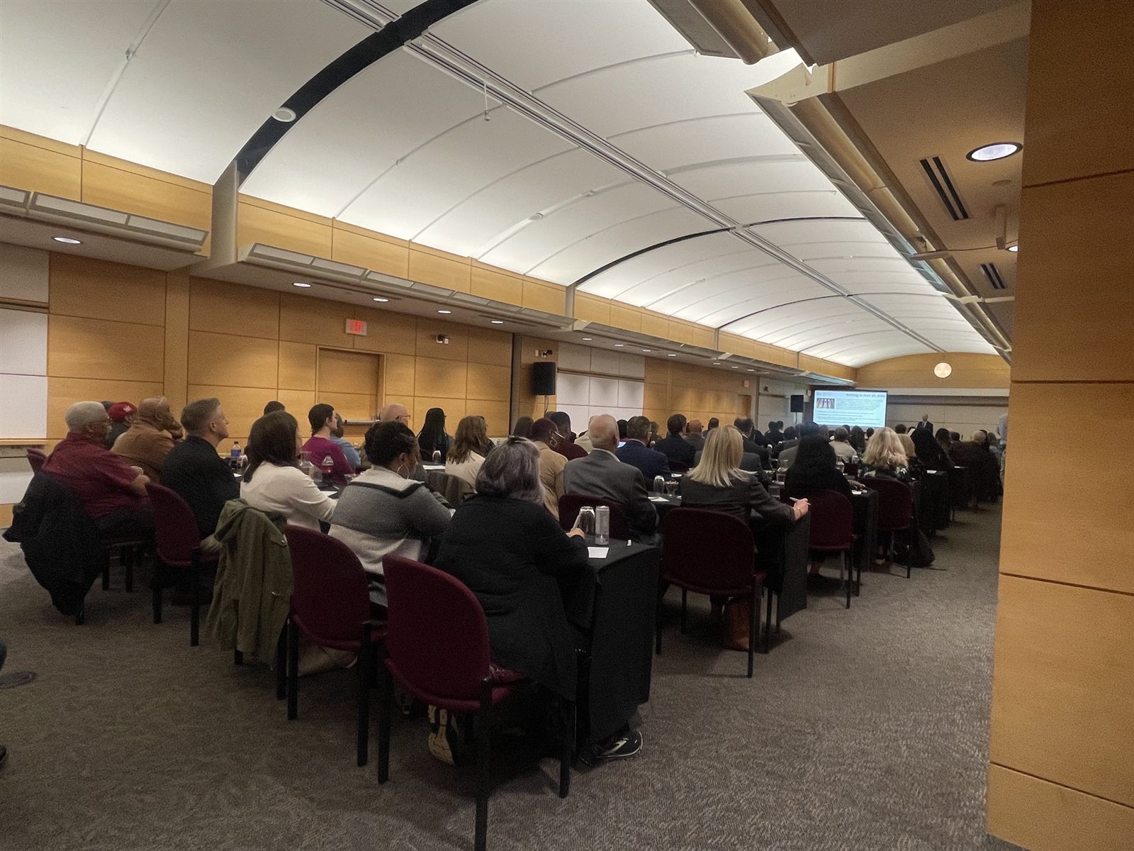 Attendees seated at rows of black-draped tables facing a presenter and projection screen in a wood-paneled conference hall