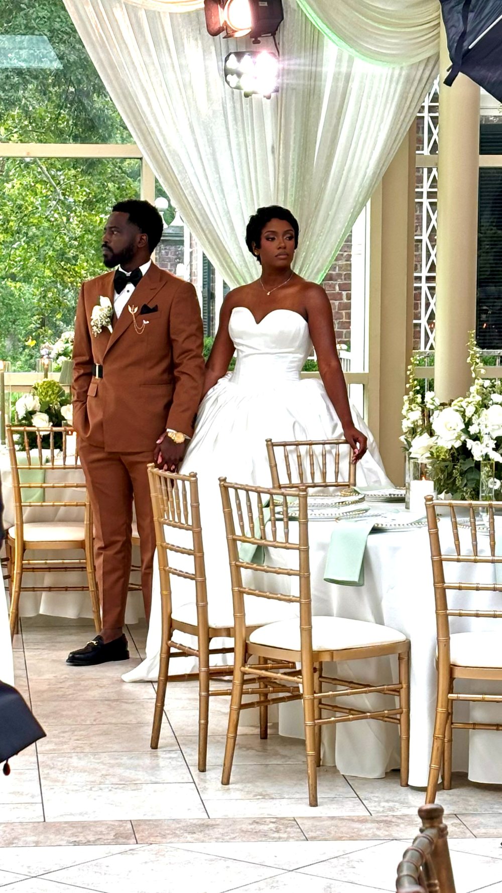 Bride in a strapless ball gown and groom in a cognac suit standing hand in hand among gold chiavari chairs at a garden reception