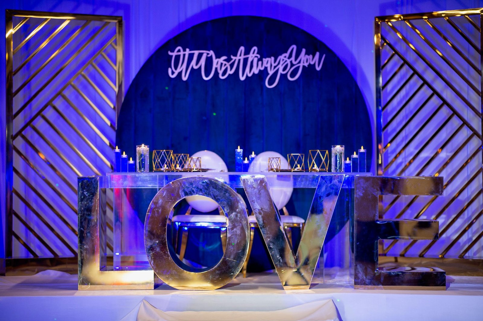 Sweetheart table behind oversized mirrored LOVE letters, with an 'It Was Always You' sign and gold geometric decor